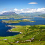 A scenic view of the Ring of Kerry, Ireland, featuring rolling emerald hills, rugged coastline, and sparkling lakes, with a winding road leading to distant mountains under a cloudy sky.